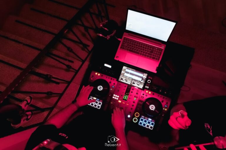 Vista dall’alto di una postazione DJ con mixer, computer e luci rosse.